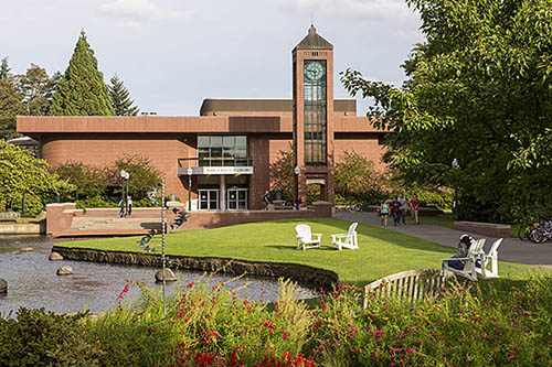 A serene image of Willamette's campus with lawn chairs near the stream and the Hatfield Library building behind