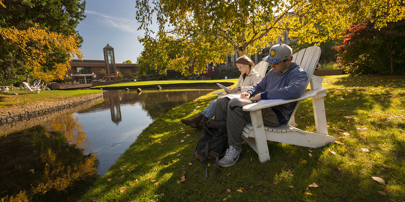 Two Willamette students sit in chairs alongside the Mill Stream at Salem campus 