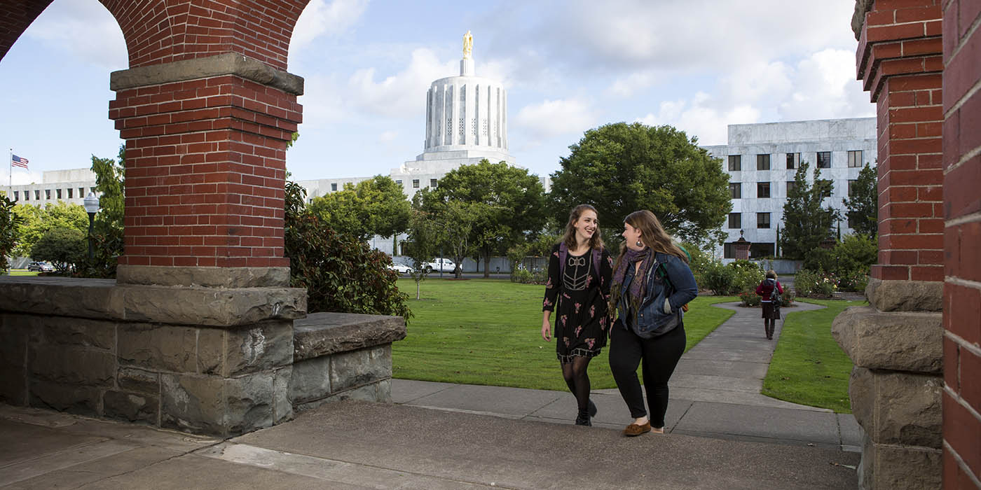 Two students walk together on campus with the state capitol building in the background, symbolizing the real-world connections and opportunities within the Politics, Policy, Law, and Ethics academic program.