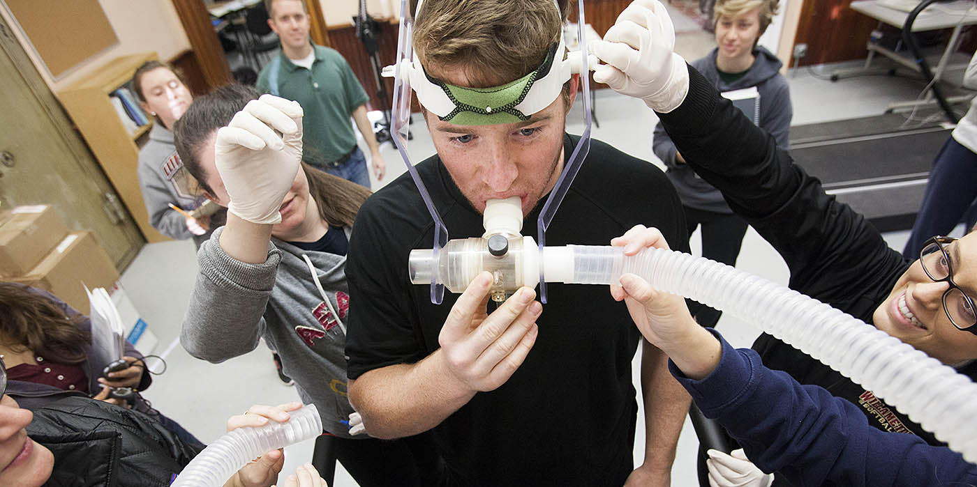 Willamette students conducting a respiratory experiment with a male participant using a breathing apparatus in a laboratory setting, assisted by several peers and overseen by an instructor.