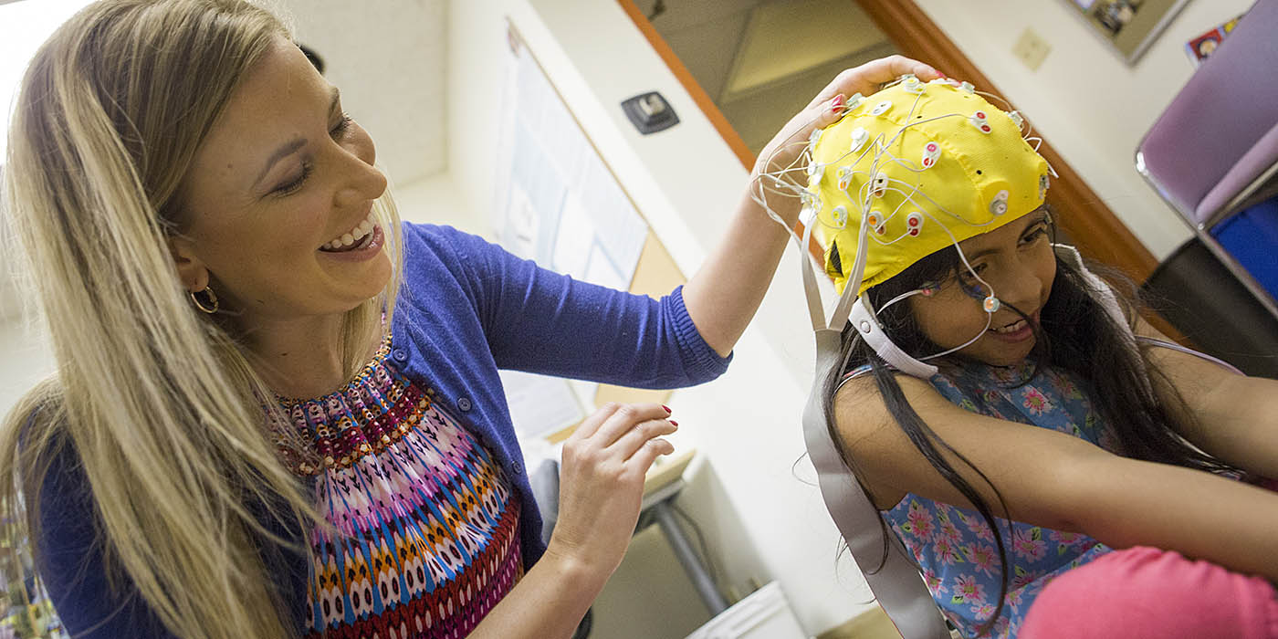 A psychology student works with a child participant wearing an EEG cap, conducting a hands-on experiment in a university research lab as part of a developmental psychology study.