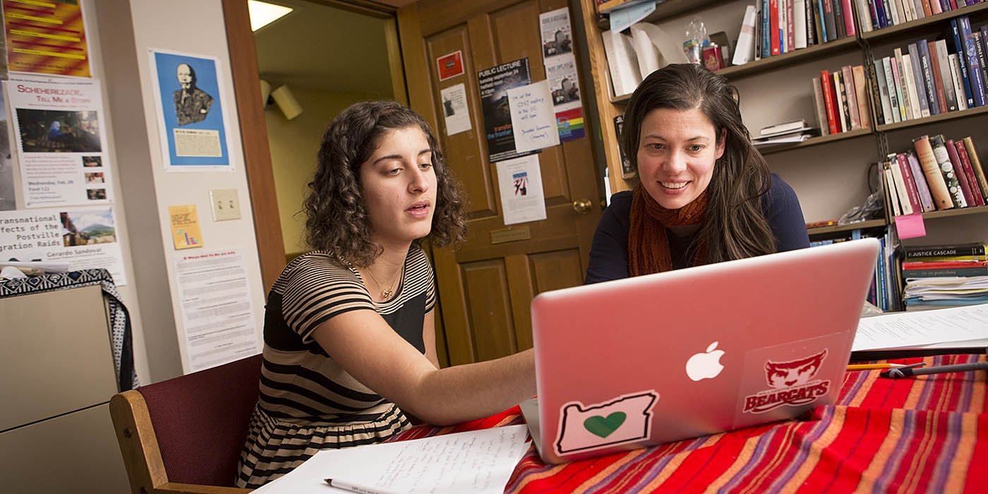 A student and professor collaborate on a project using a laptop in a Women's and Gender Studies academic office, surrounded by books and posters, reflecting the program's focus on critical thinking and academic engagement.