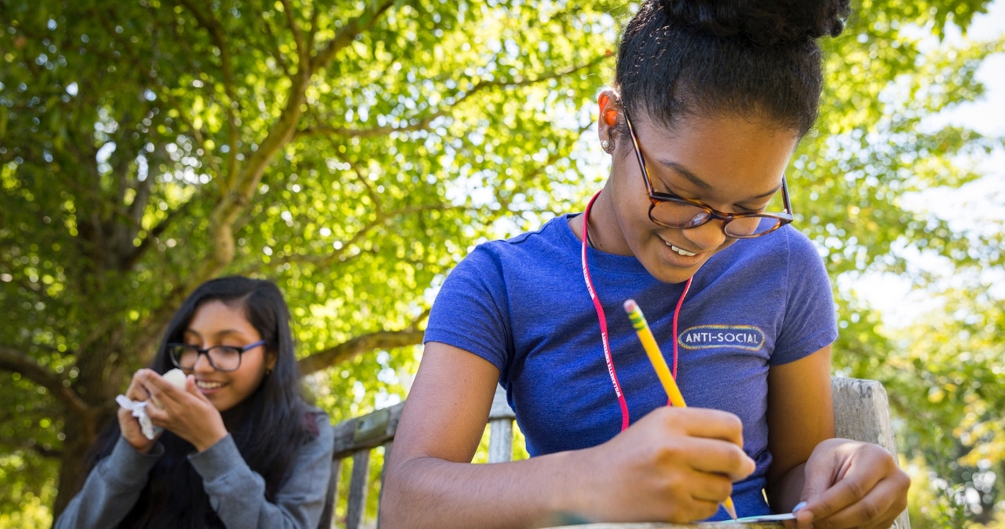 Two students with the first one writing something down while both are sitting in nature
