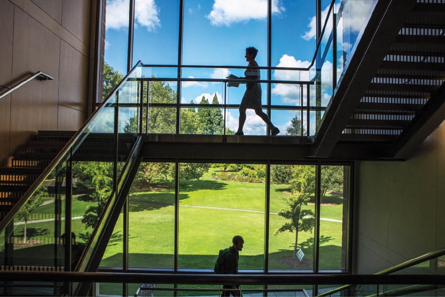 A teacher and a student walking around stairs that overlook windows and a green space