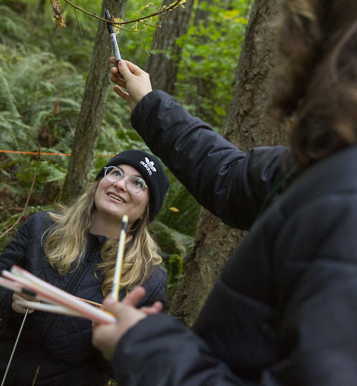 Two female students conduct field research in a forest, one holding up a branch with a sharpie while the other observes 