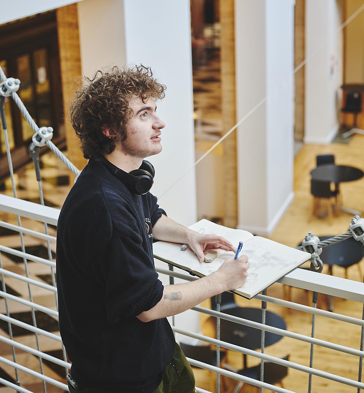 Student sketches in a notebook while observing an indoor space from an indoor balcony.