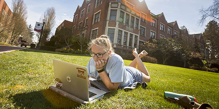 A student laying in the lawn looking at a computer
