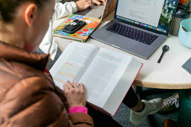 Student reviewing a book in front of a laptop.